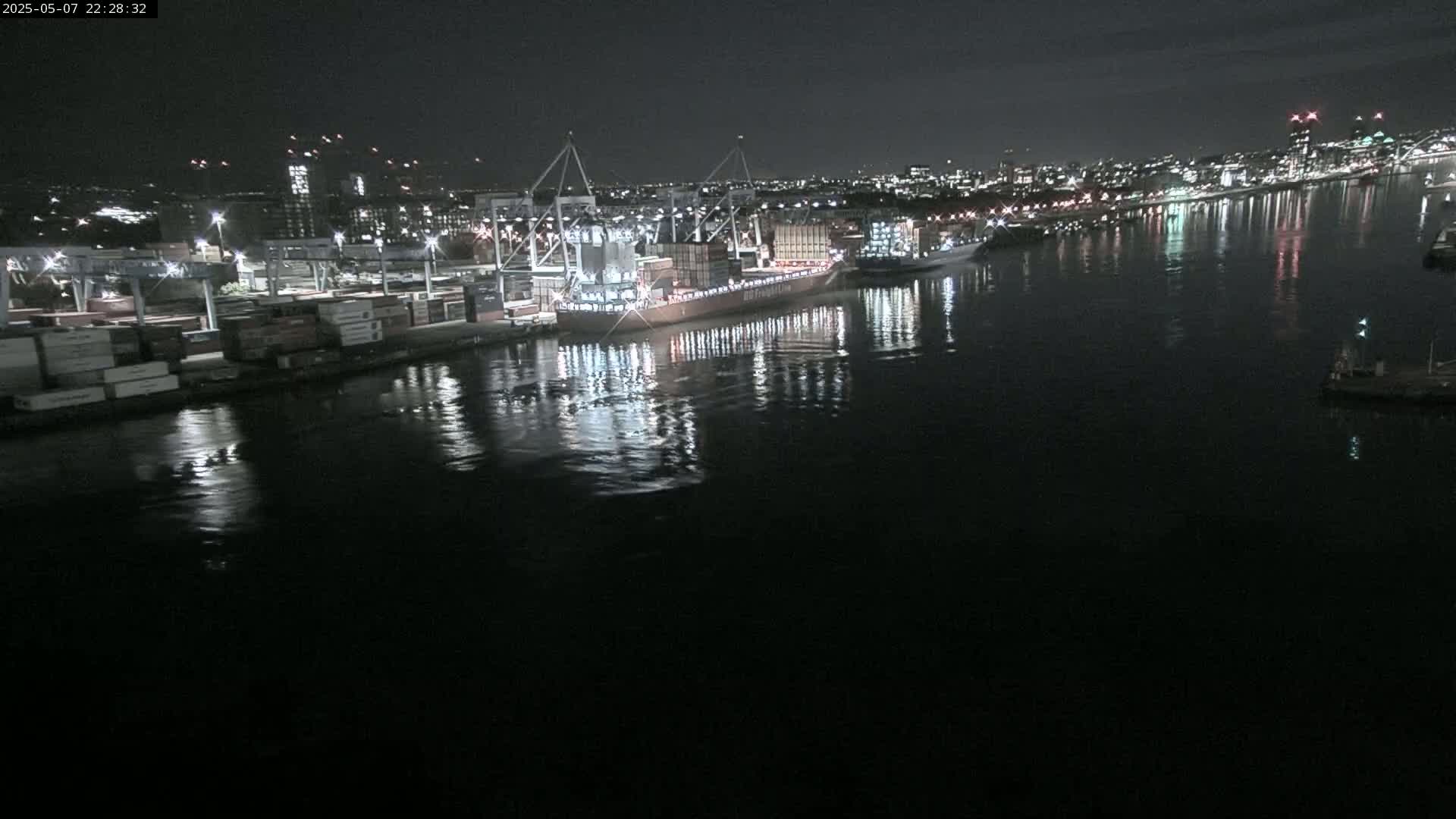 A large container ship is docked at a nighttime port, its lights reflecting on the calm water.