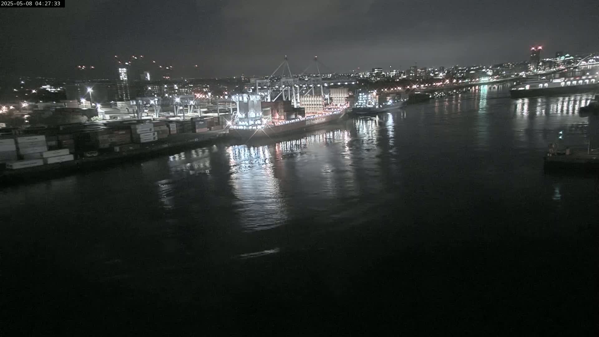 A large container ship is docked at a nighttime port, with smaller vessels nearby and city lights in the background under a dark, cloudy sky.