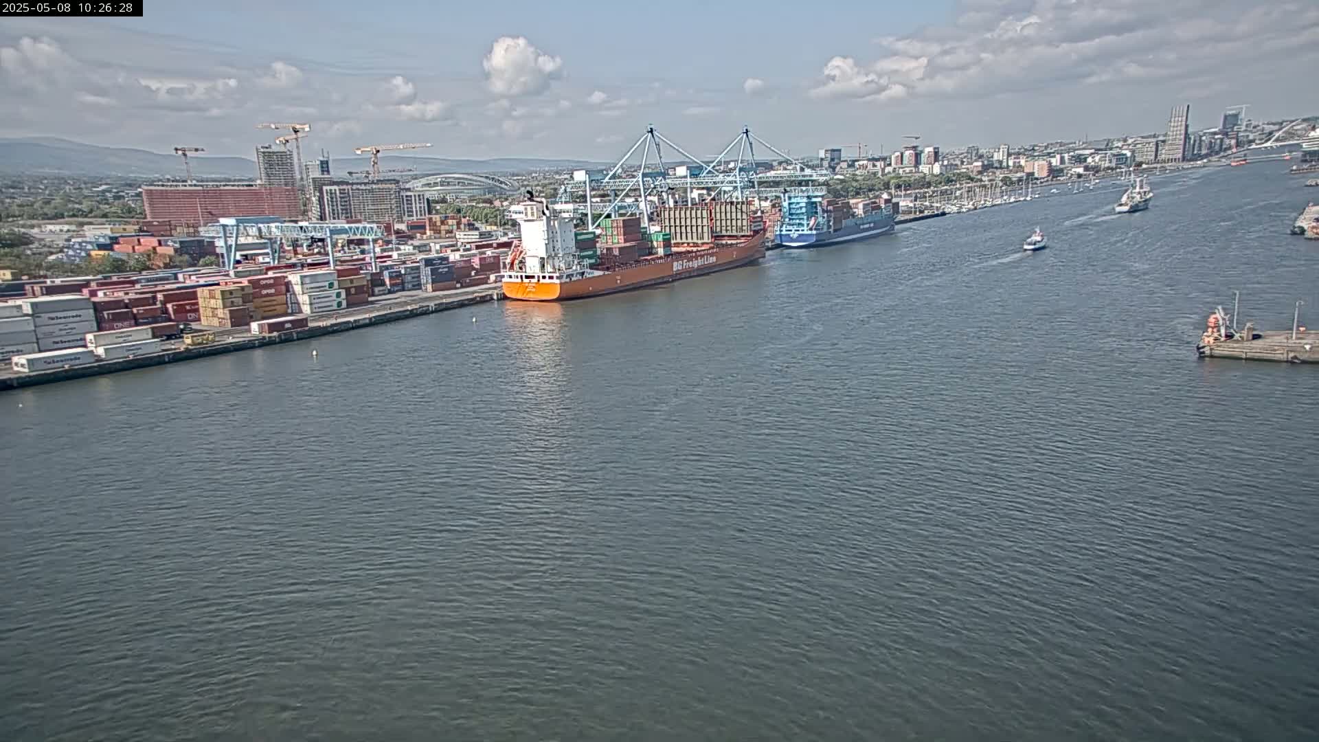 A container ship is docked at a port on a partly cloudy day, with other ships in the water and a city skyline in the background.