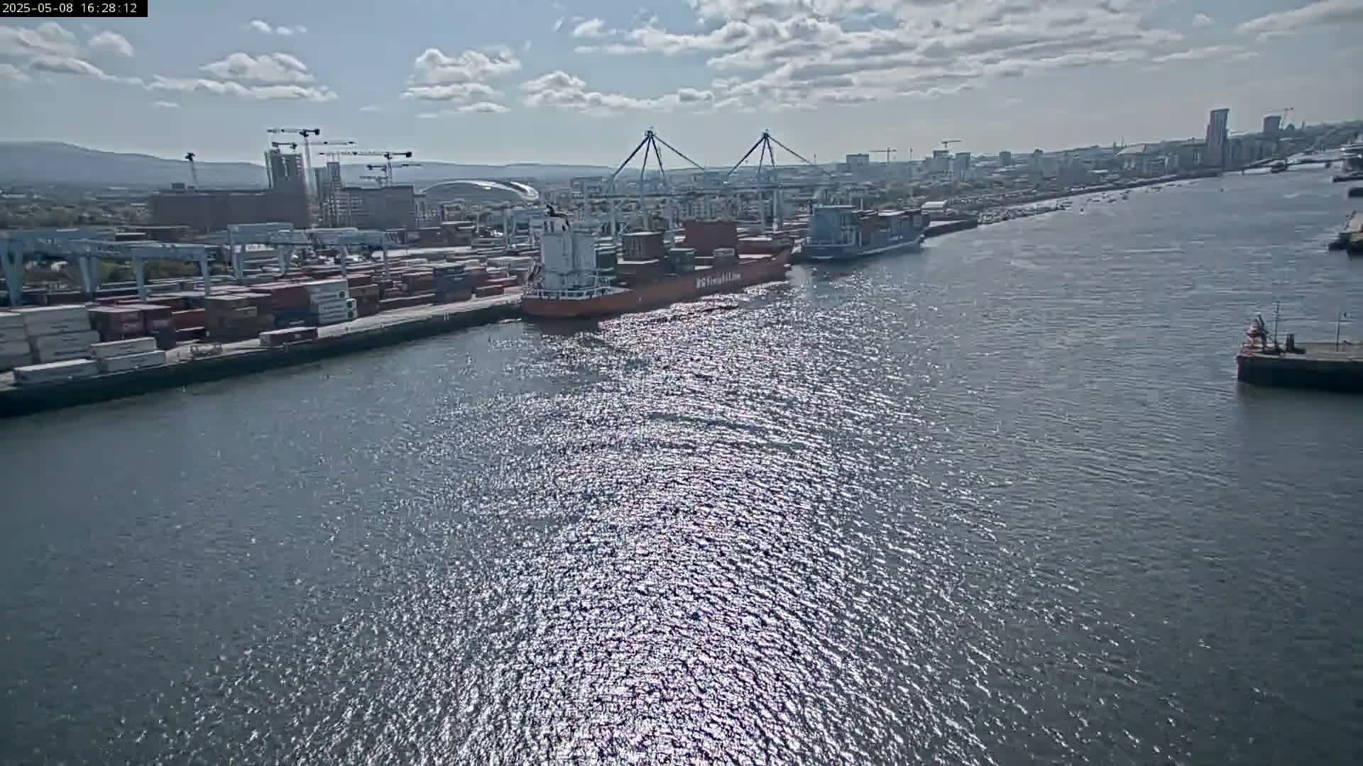 A container ship and other vessels are docked at a busy port on a partly sunny day.