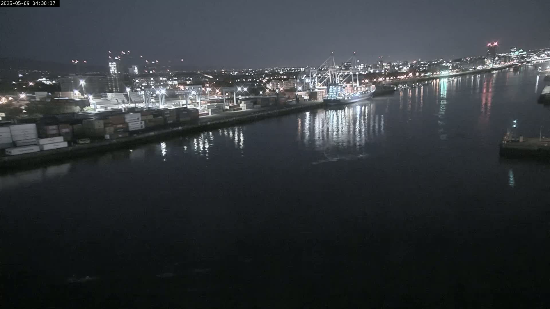A nighttime aerial view of a harbor shows a cargo ship and numerous containers at a dock, with city lights reflecting on dark water.