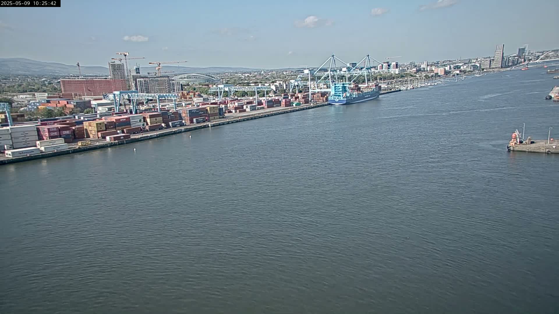 A container ship is docked at a busy port on a calm, sunny day.