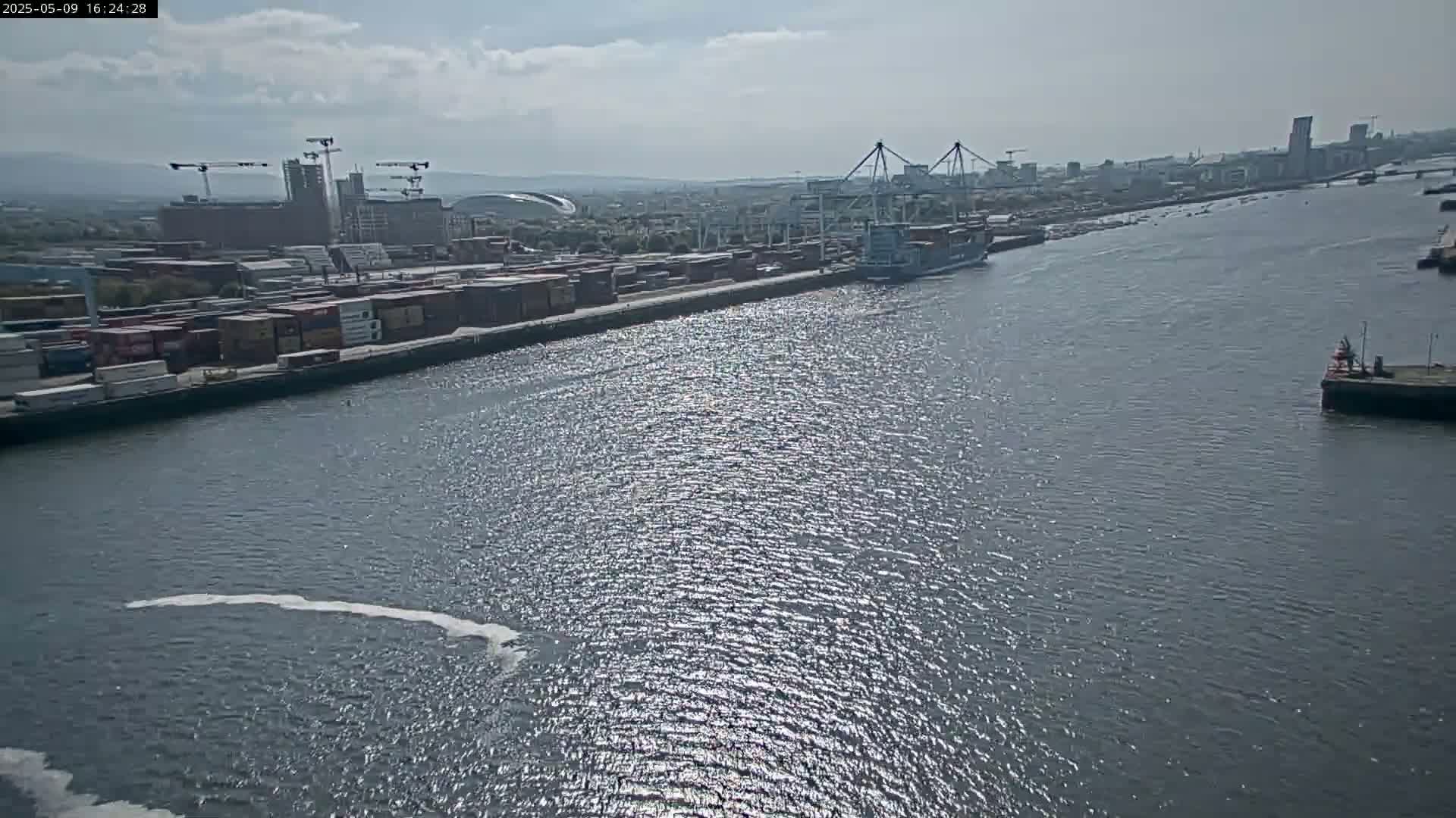 A container ship is docked at a port on a sunny day, with numerous shipping containers visible along the waterfront.