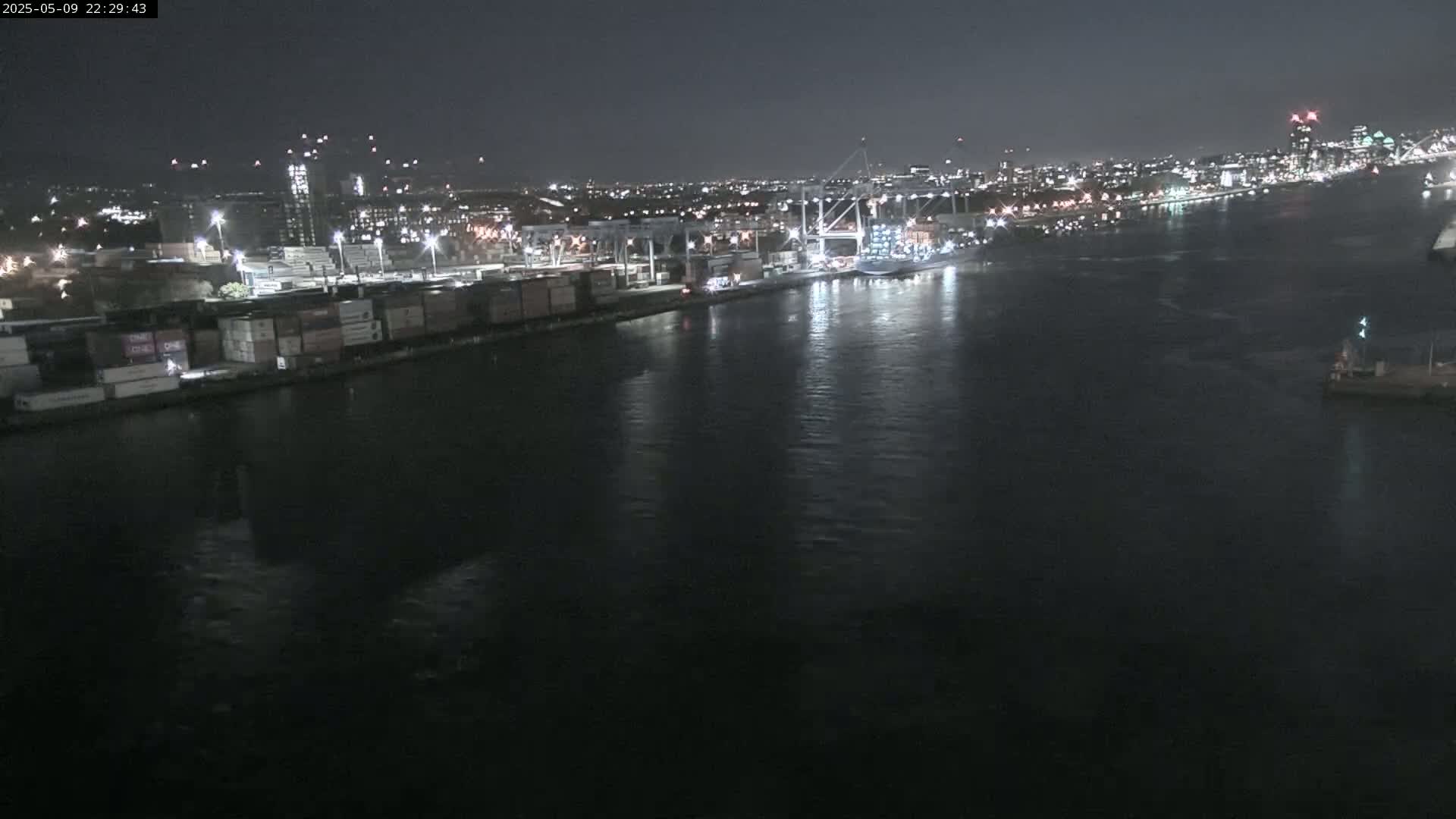 A nighttime view of a shipping port with numerous cargo containers and cranes along a dark, calm body of water, with a city skyline visible in the background.
