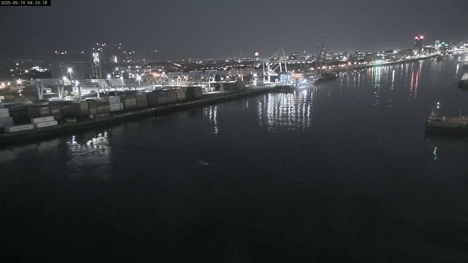 A nighttime view of a harbor with cargo containers along a dock, illuminated by lights, reflecting on dark water, under a mostly clear sky.