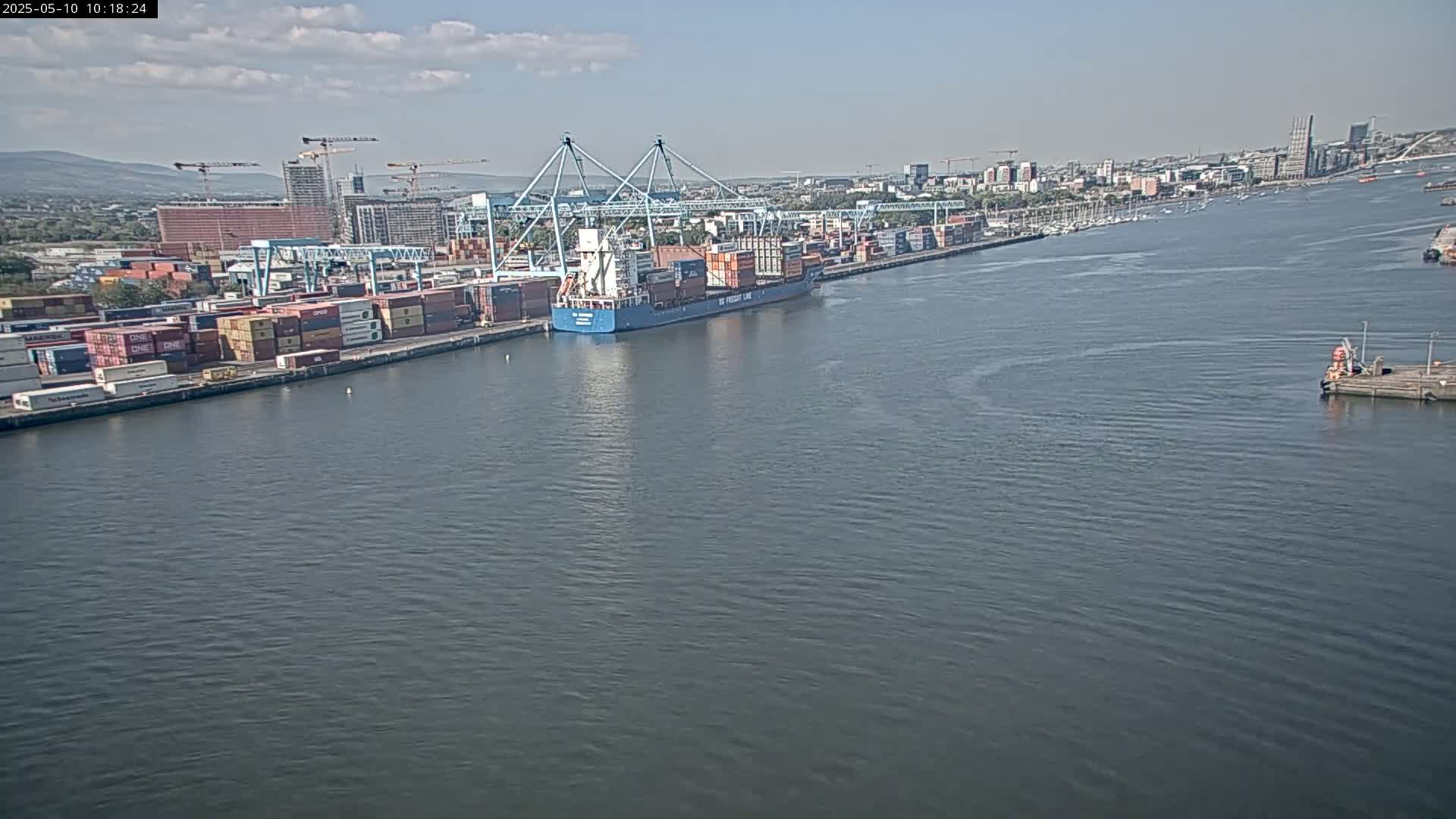A container ship is docked at a busy port on a partly cloudy day, with a city skyline visible in the background.