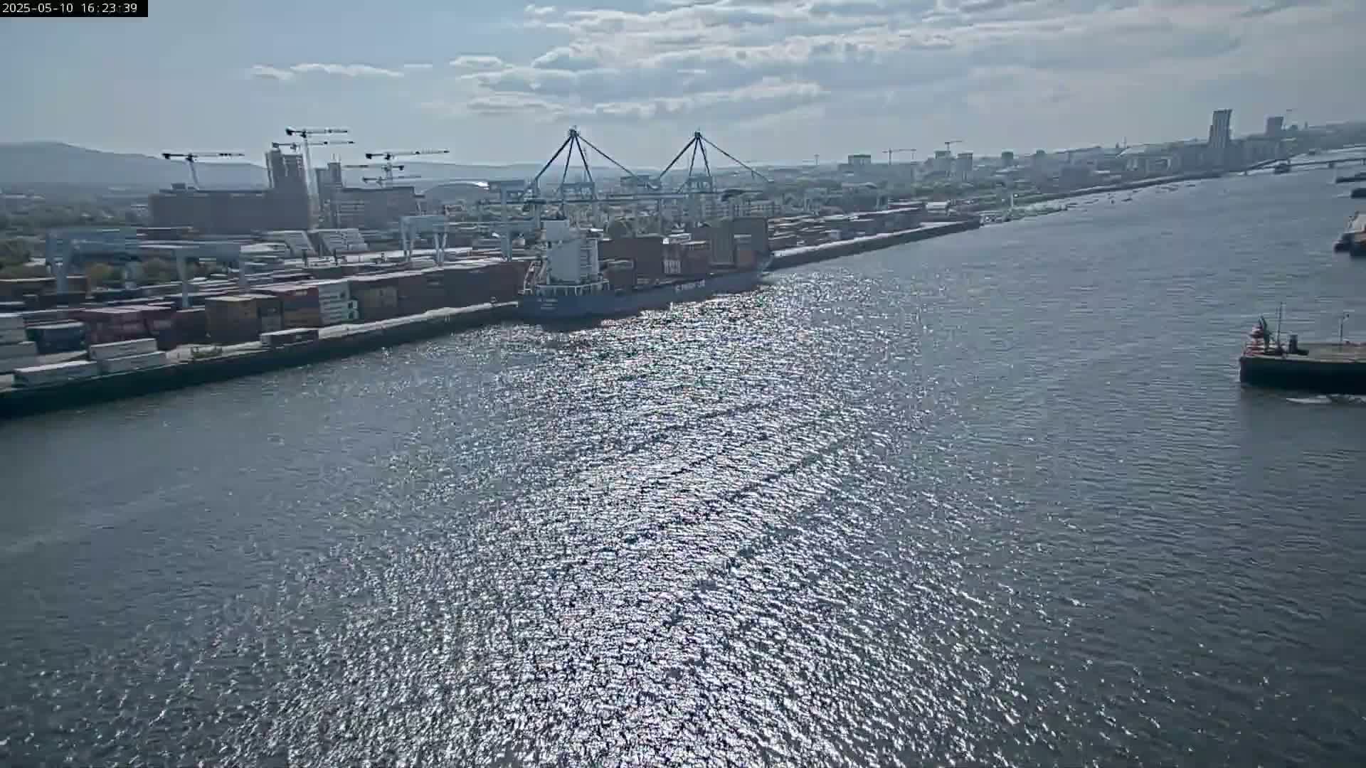 A container ship departs a port on a partly sunny day, passing by numerous cargo containers and cranes along a waterway.
