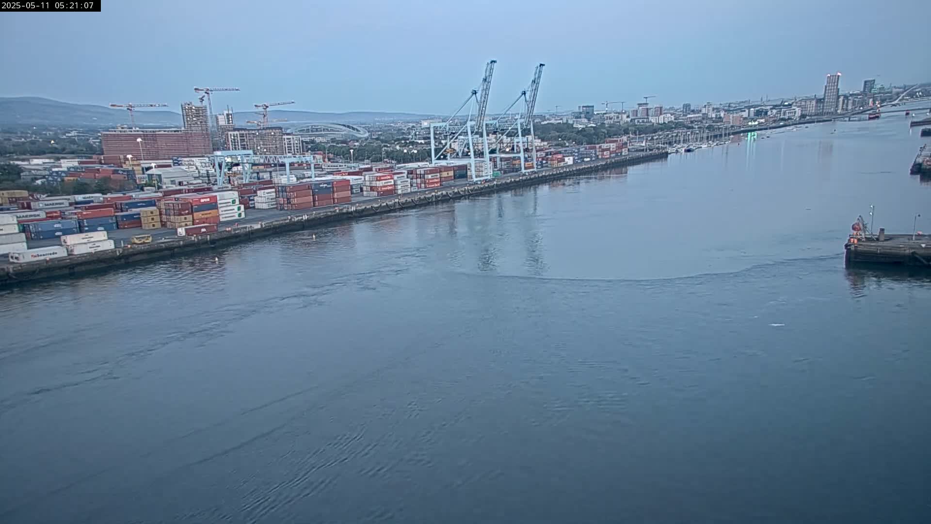 A container port with several cranes and many shipping containers is situated on a body of water under a cloudy sky.