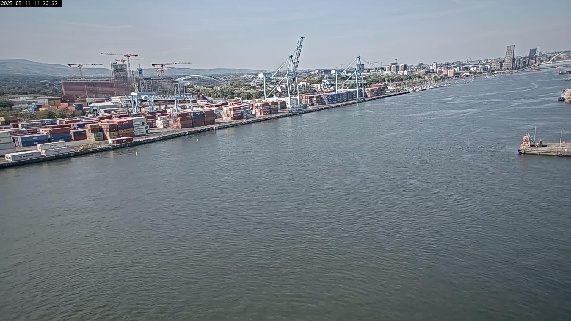 A container port with numerous cargo containers and cranes sits on a body of water under a clear sky.