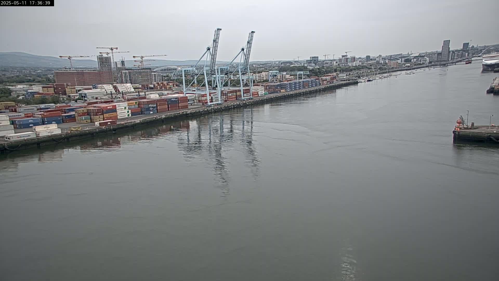 A large container port with several cranes is visible along a calm, gray waterway under an overcast sky.