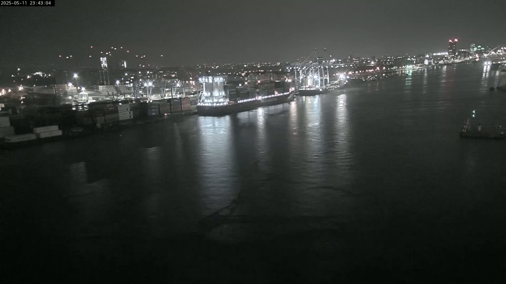 At night, a large container ship and other vessels are docked at a busy port, illuminated against a dark, calm body of water.