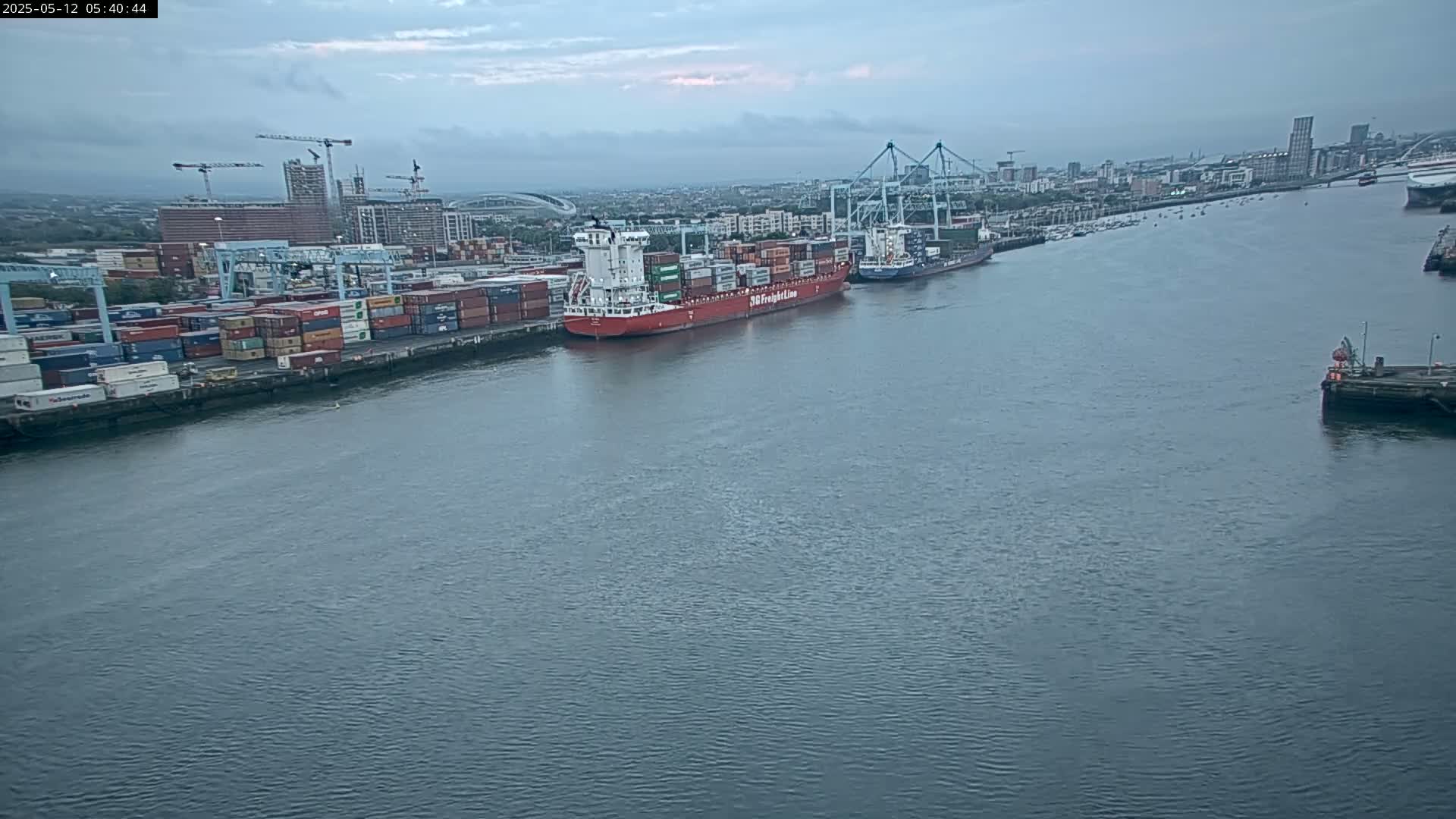 A large container ship is docked at a busy port under an overcast sky.