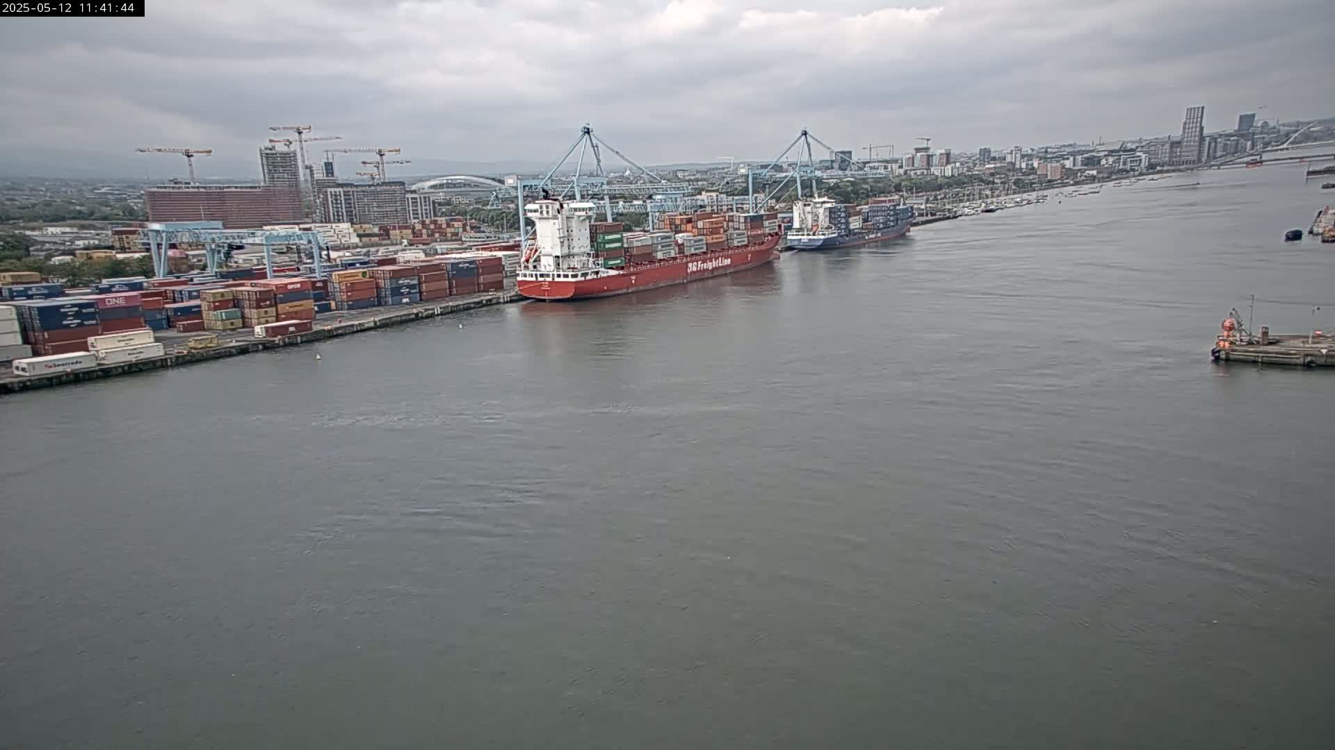 A large container ship and several smaller vessels are docked at a port under an overcast sky.