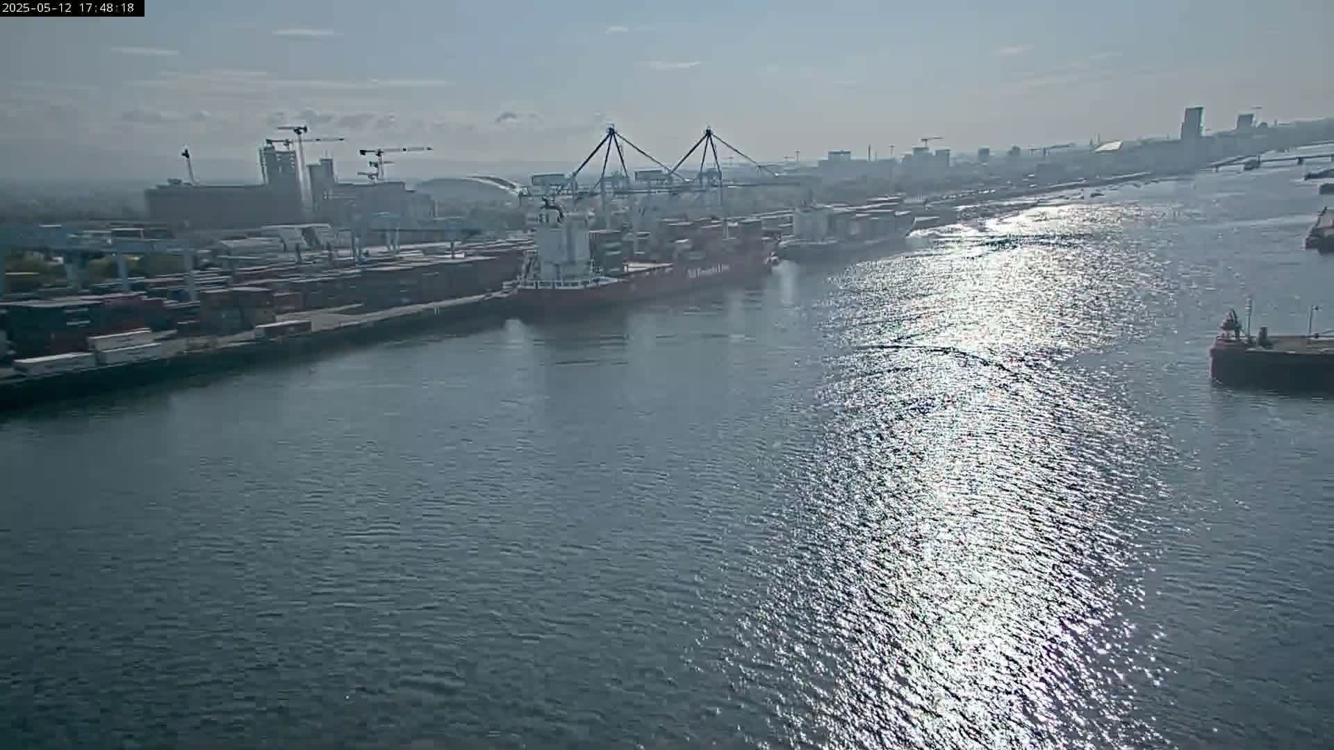 A container ship and barge are docked at a port on a sunny day, with other ships and buildings visible in the distance across the waterway.