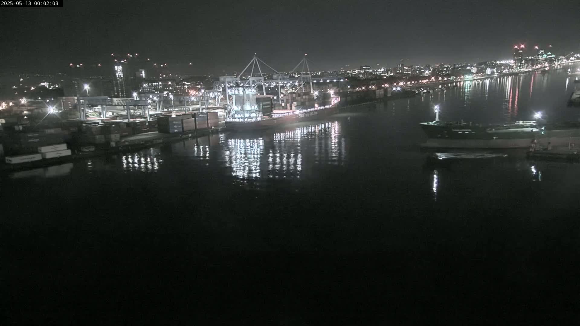 At night, a large container ship is docked at a port, with other smaller vessels nearby, reflecting in calm water under a dark sky.