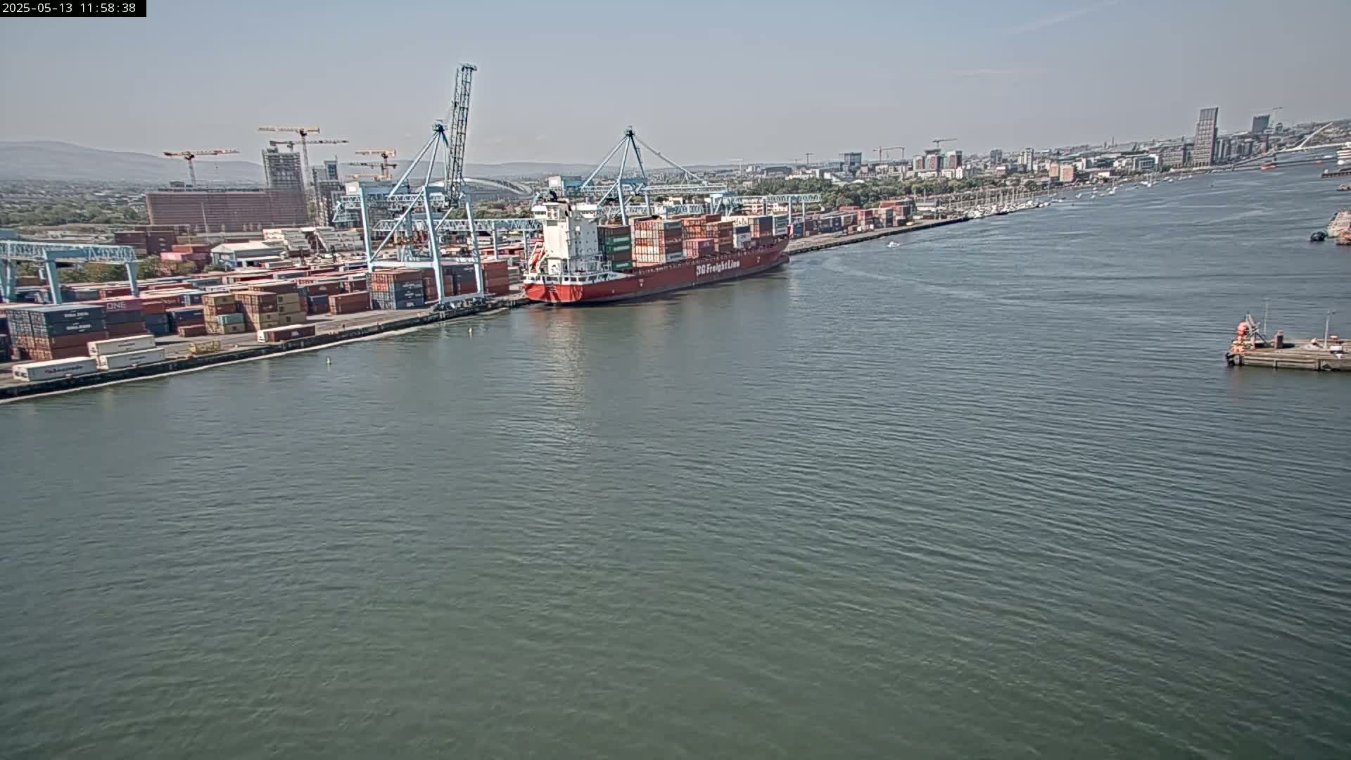 A large container ship is docked at a port on a calm, sunny day, with numerous containers visible on the dock and in the background.