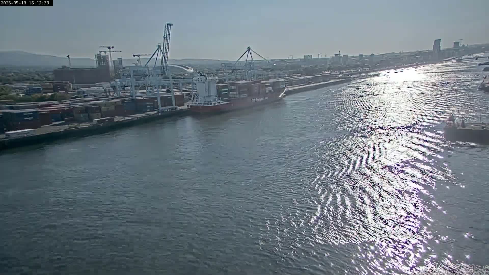 A container ship is docked at a port on a sunny day, with its cargo containers visible near multiple cranes and other industrial structures.