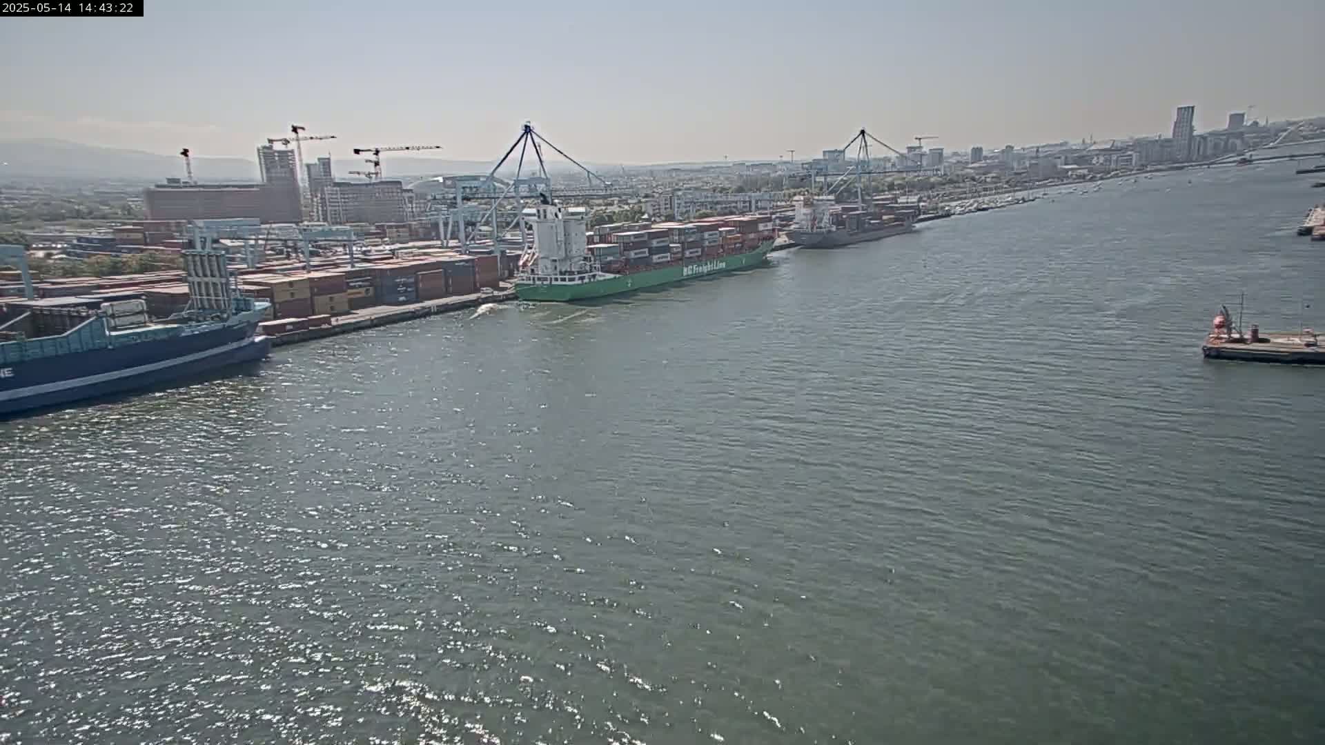 A container ship and other vessels are docked at a port on a calm, sunny day.