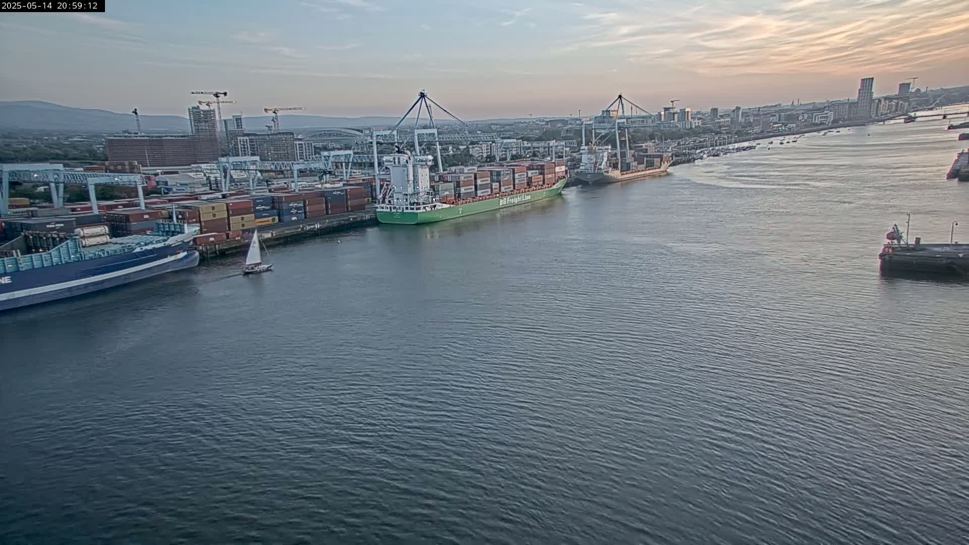 A container ship and other vessels are docked at a port under a partly cloudy evening sky.