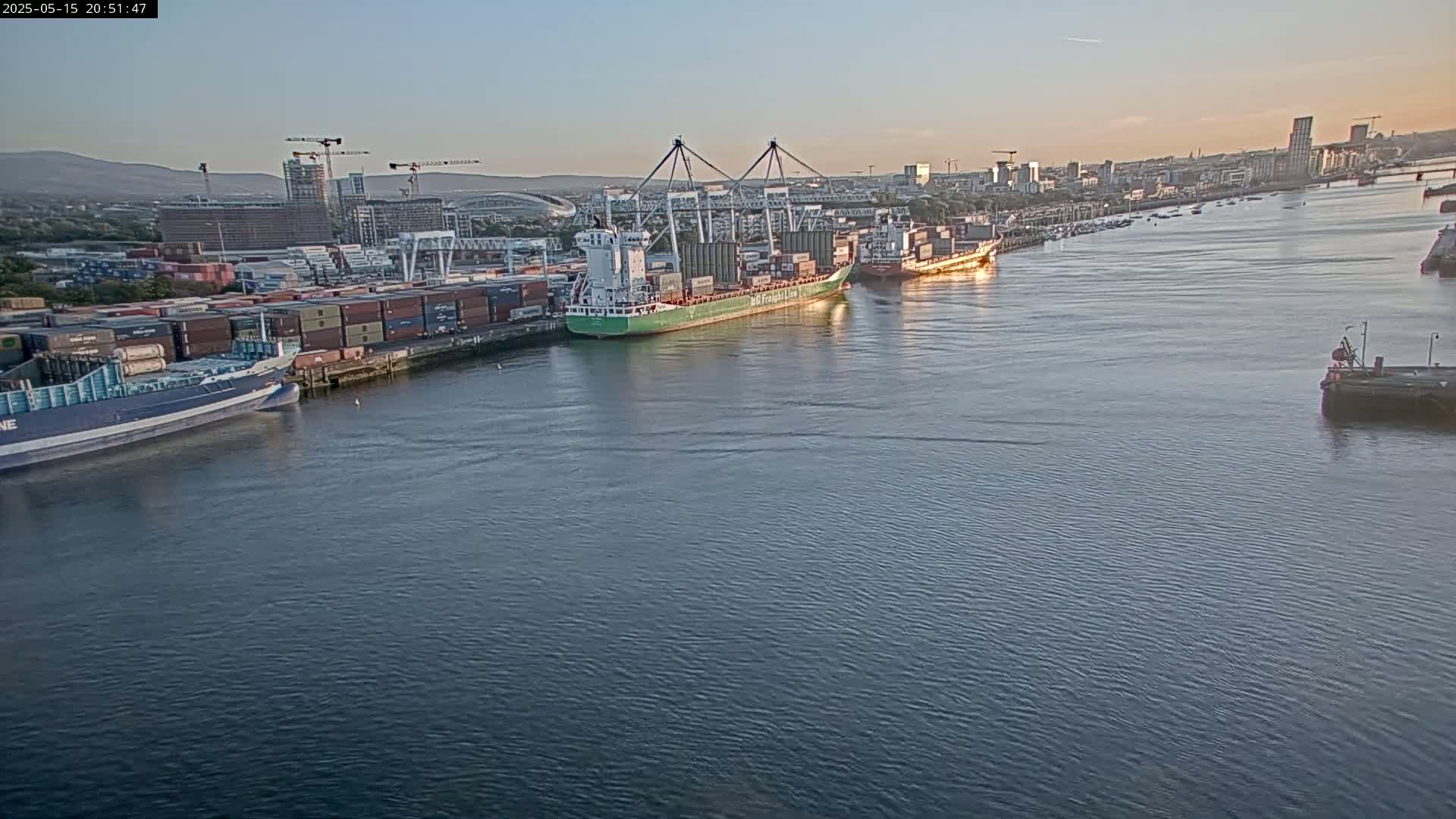 Several container ships are docked at a port in calm evening water, with a city skyline visible in the background.