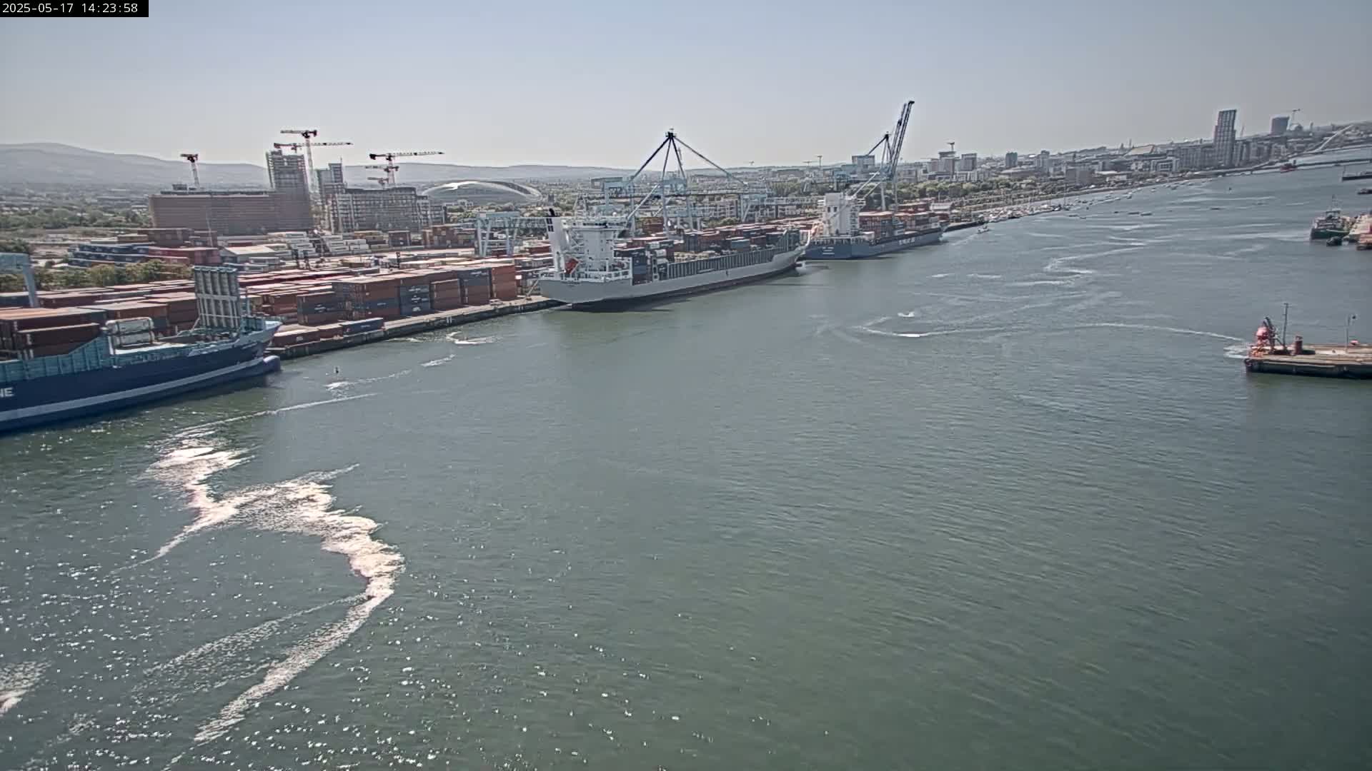 A container ship is docked at a busy port on a calm, sunny day, with other ships and a cityscape visible in the background.