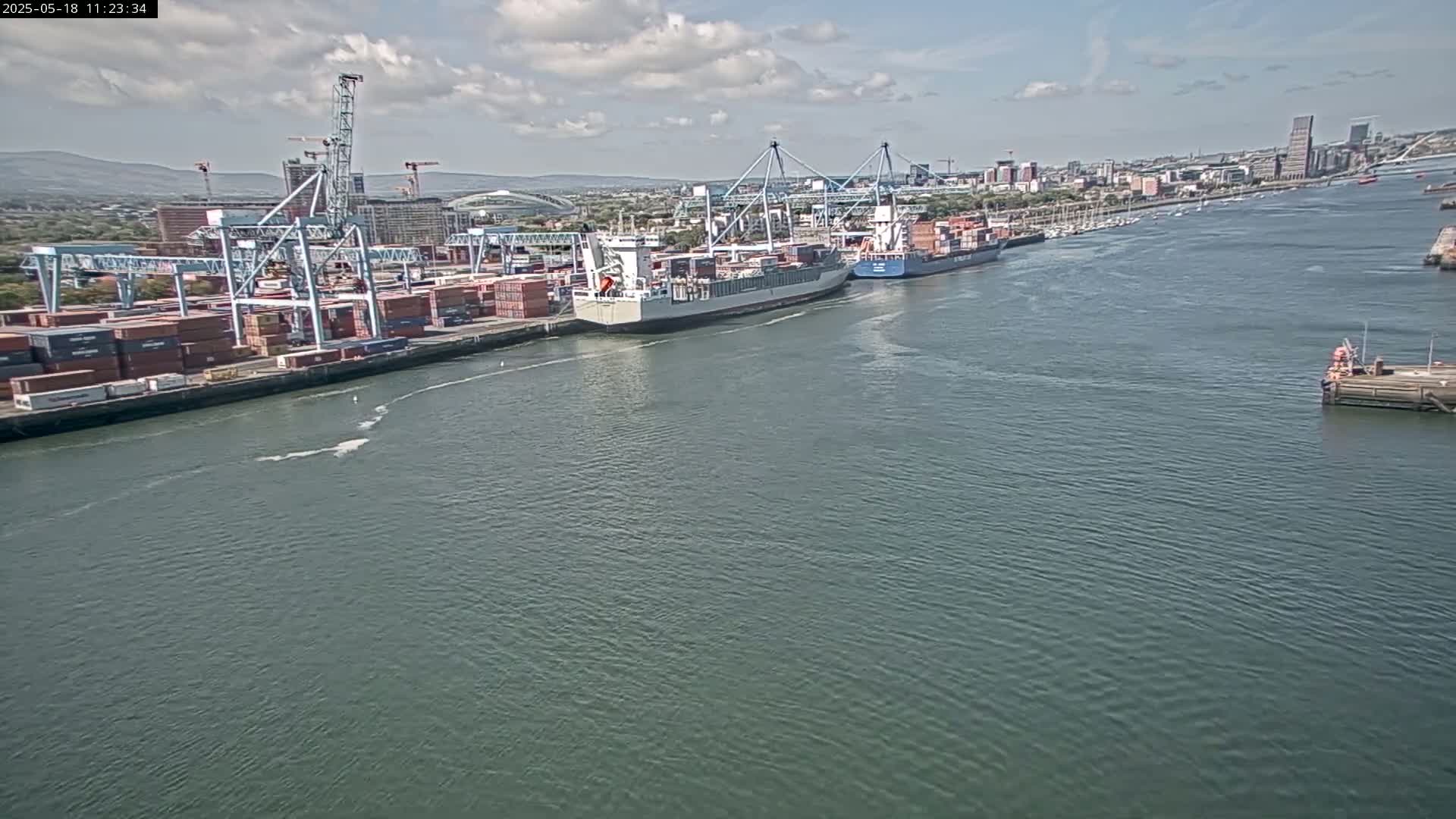 A container ship is docked at a busy port on a partly cloudy day.
