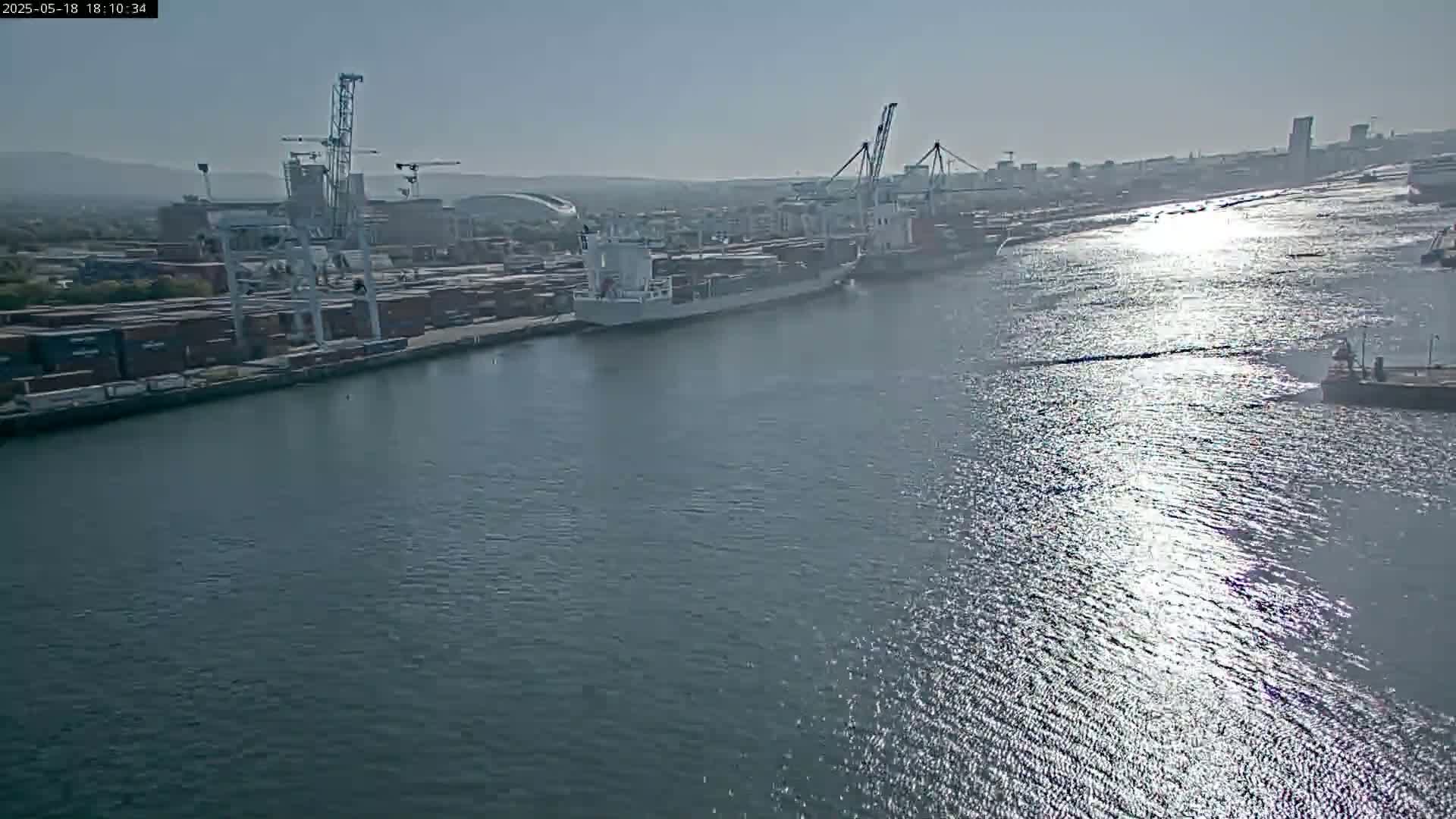A large cargo ship is docked at a busy port on a sunny day, with numerous shipping containers and cranes visible along the waterfront.