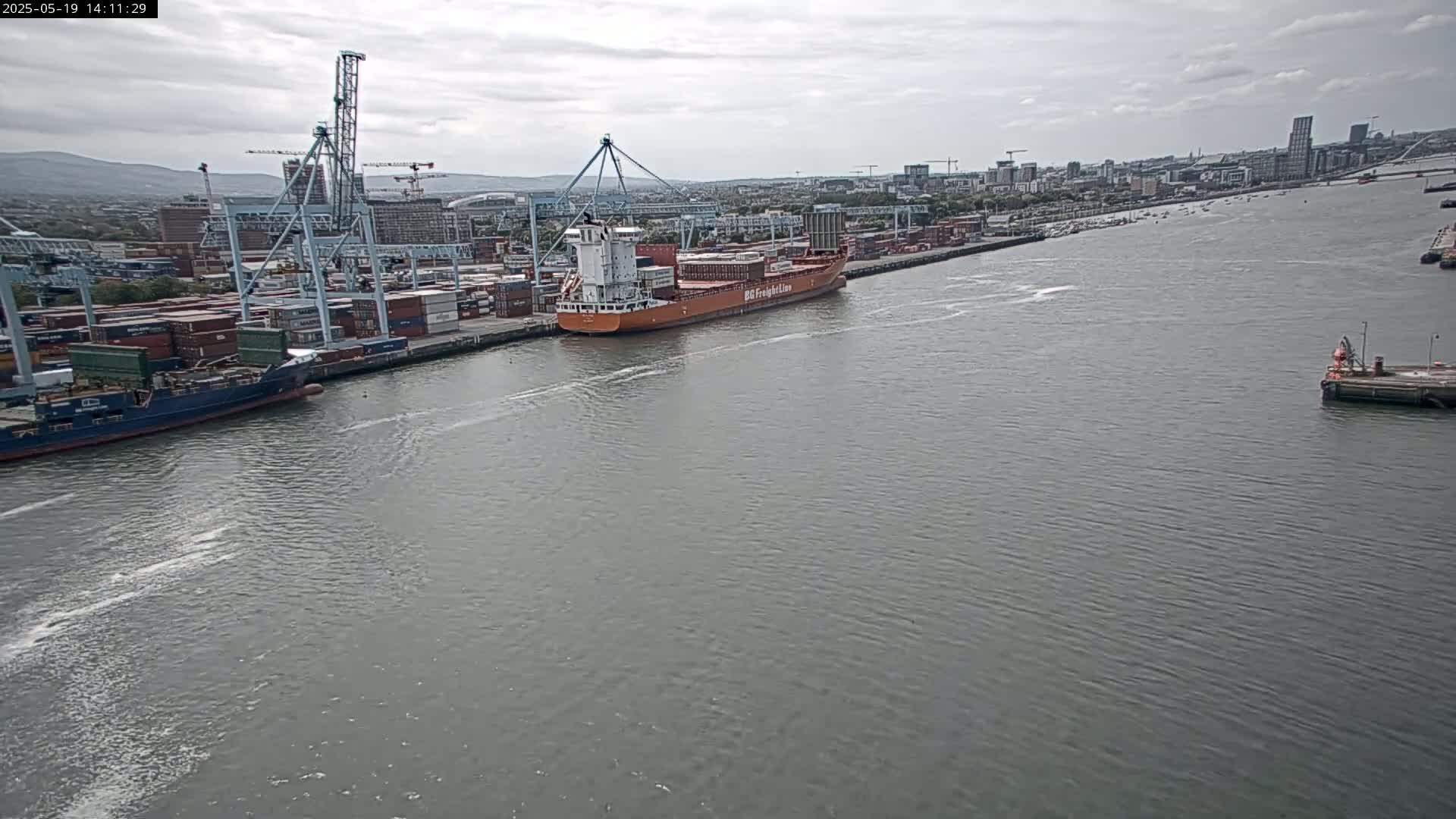 A large container ship is docked at a busy port under overcast skies, with several other ships and cranes visible.