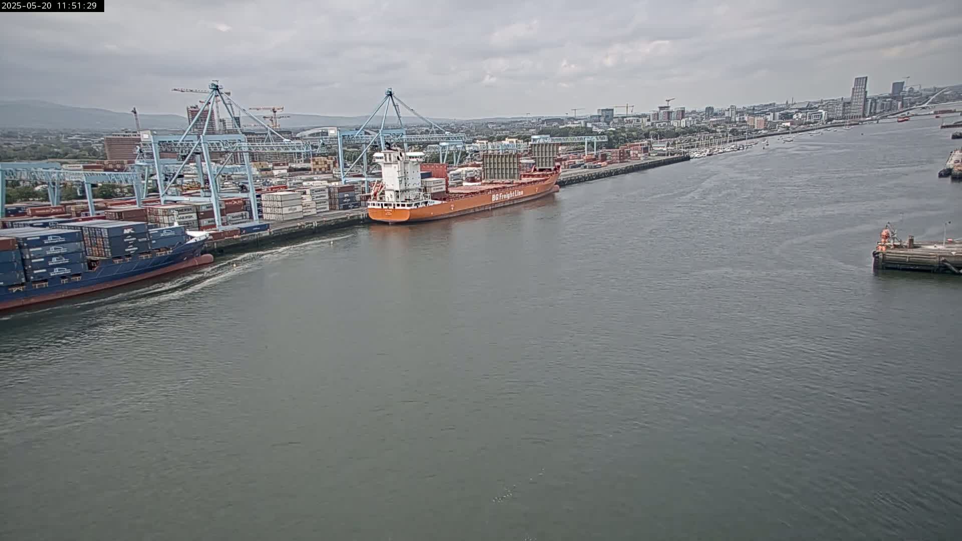 Two container ships are docked at a port, under an overcast sky, with a city skyline visible in the background.