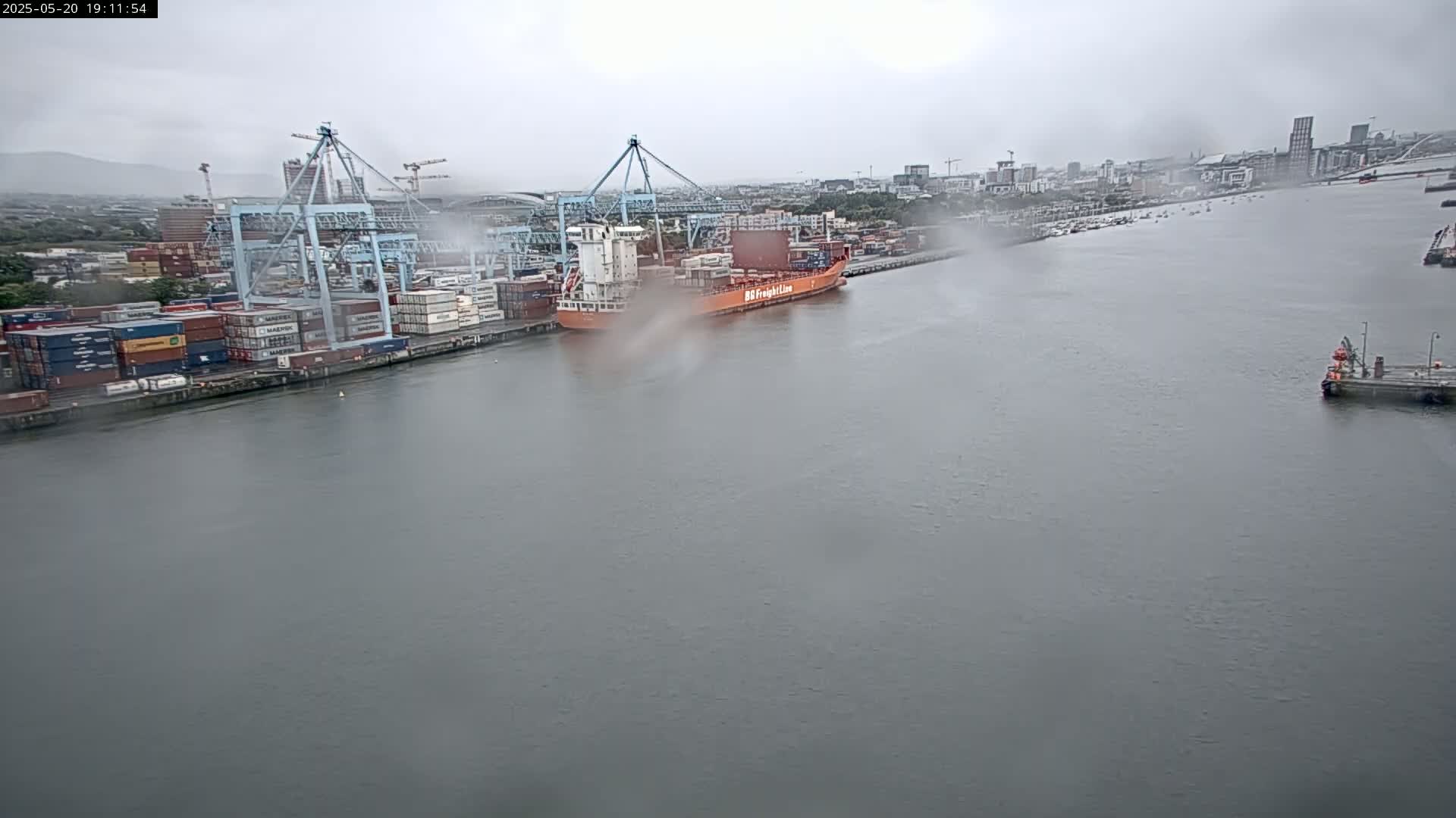 A large container ship is docked at a port, surrounded by numerous shipping containers and cranes under an overcast sky.