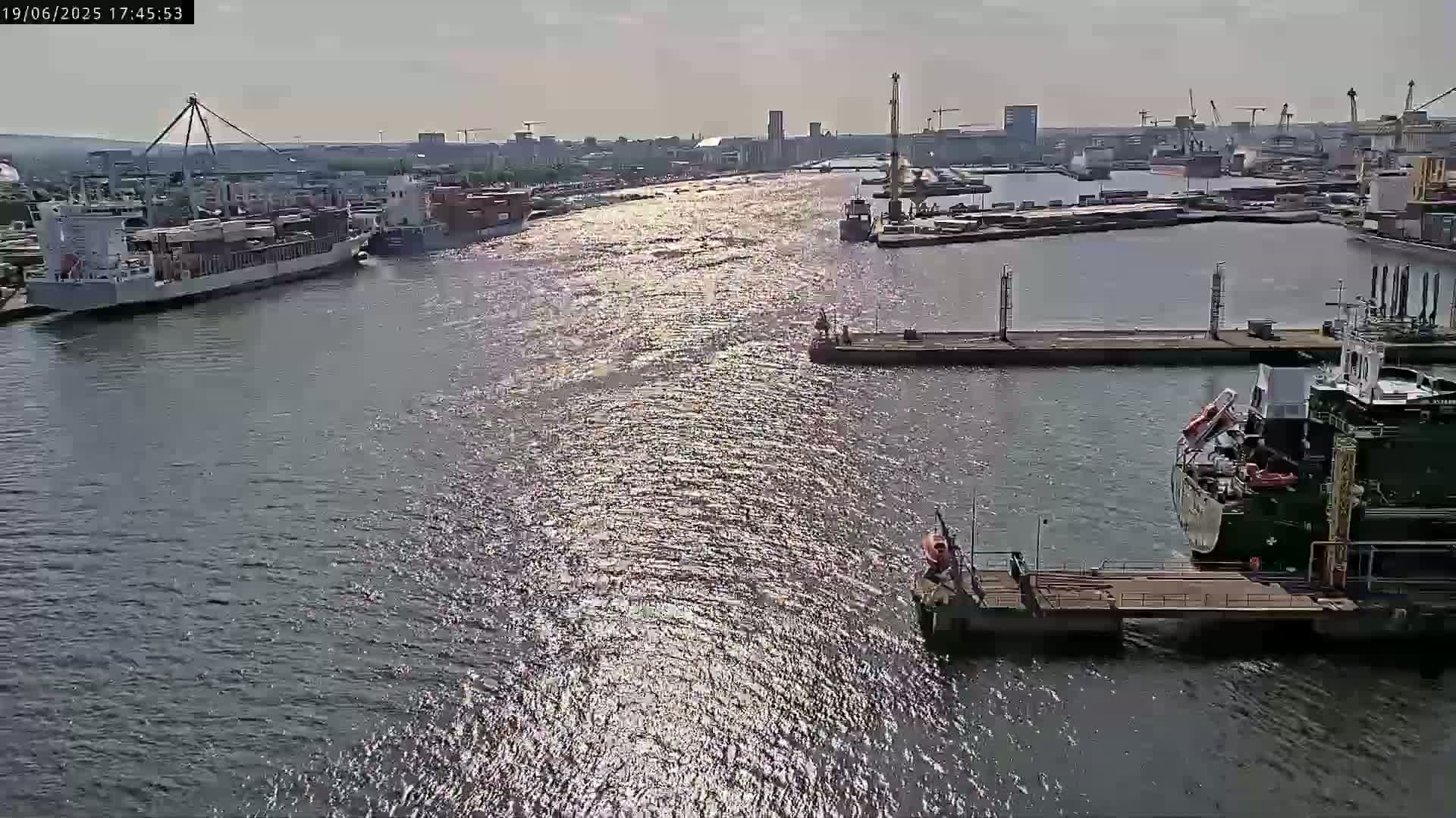 A sunny day at a harbor shows several ships docked and in the waterway, with sparkling water reflecting sunlight.