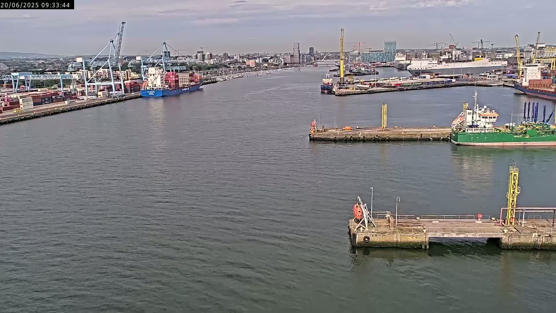 A wide shot of a harbor on a partly cloudy day shows several ships and docks, with a container ship prominently featured near the left side.