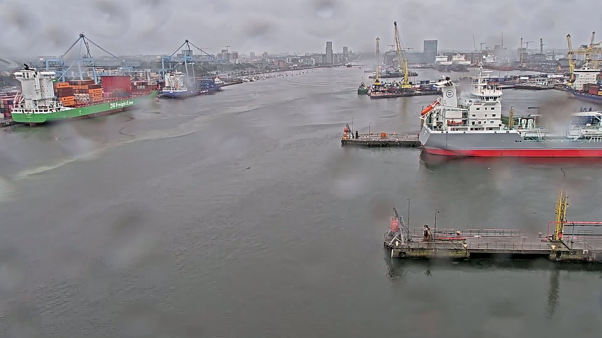 Several cargo ships and tankers are moored in a harbor on a rainy day.