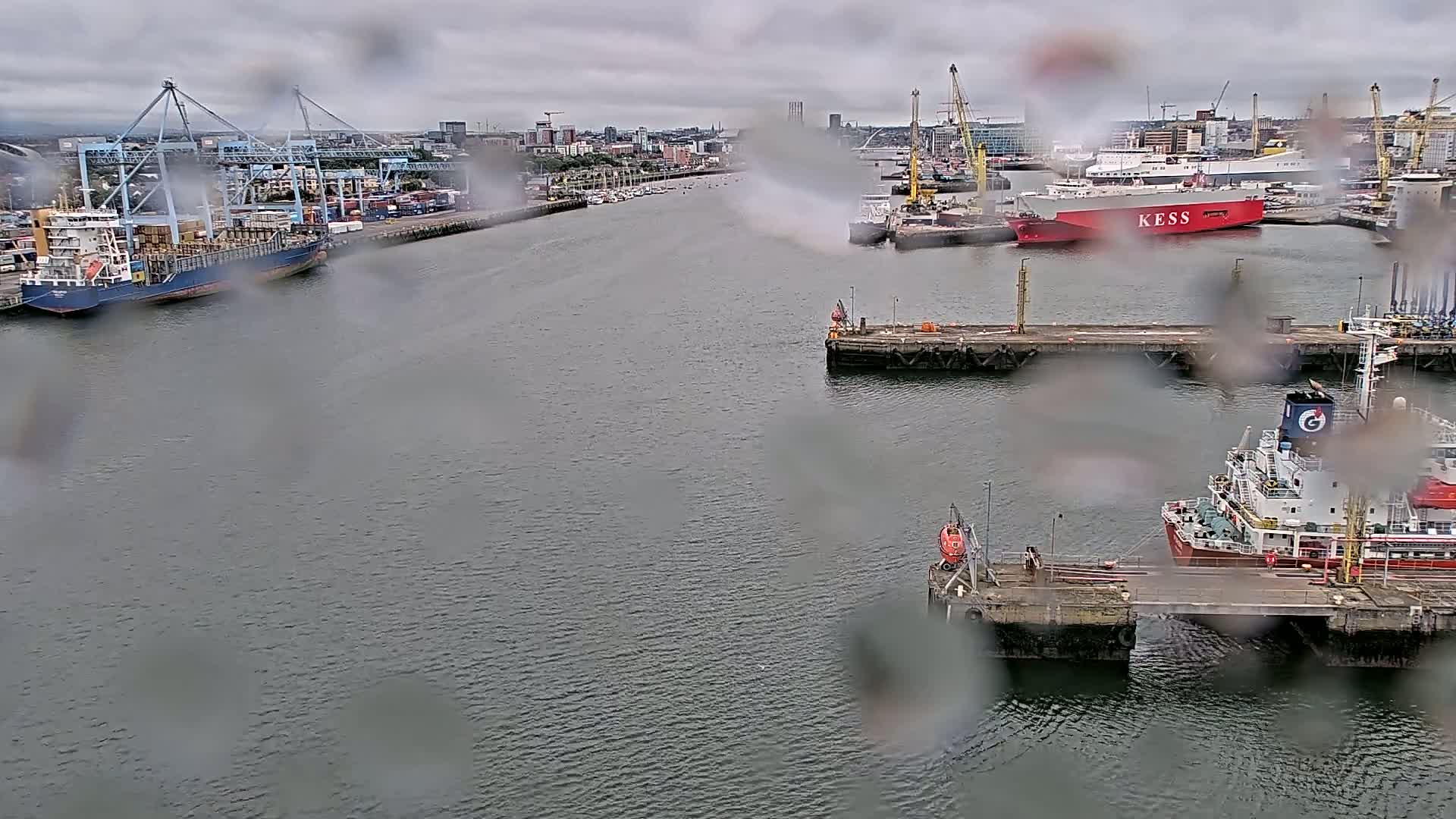 A harbor scene on an overcast day, viewed through rain-covered glass, shows several cargo ships and tankers moored at docks, with cranes and other port infrastructure visible in the background.