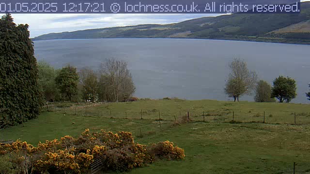 A grassy field with a wire fence overlooks a large, calm lake surrounded by mountains under a cloudy sky.