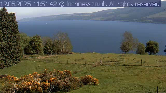 A grassy field with a low fence overlooks a large, dark blue lake nestled between rolling hills under a mostly sunny sky.