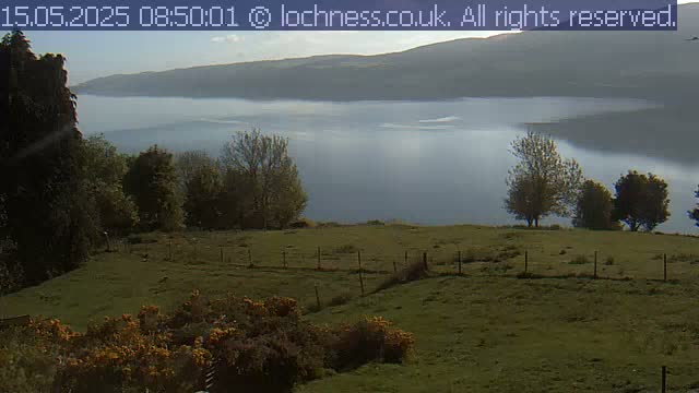 A grassy field with a low wire fence overlooks a misty loch and distant hills under a partly cloudy sky.