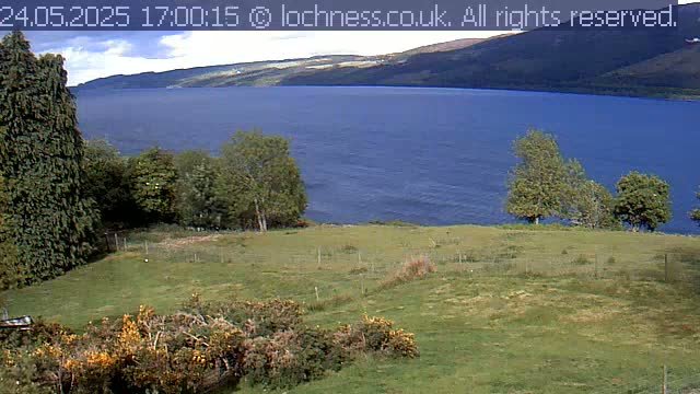 A grassy field slopes down to a large, calm lake nestled between rolling hills under a partly cloudy sky.