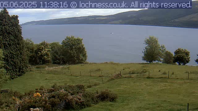 A grassy field with a short fence overlooks a calm lake and distant mountains under a partly cloudy sky.