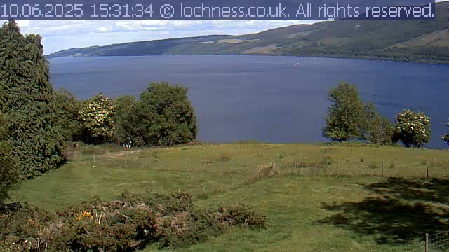 A grassy hillside overlooks a calm, blue lake nestled between rolling green hills under a partly cloudy sky.