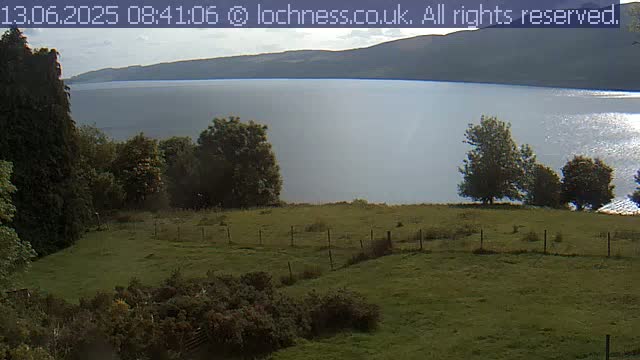 A grassy field with a low wire fence overlooks a calm lake and distant mountains under a partly cloudy sky.