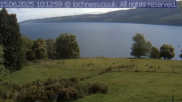 A grassy field with a wire fence overlooks a calm lake and distant mountains under a partly cloudy sky.