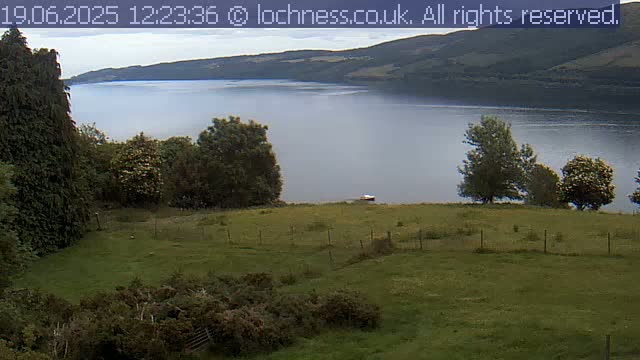 A grassy field slopes down to a calm lake, surrounded by dark green trees and hills under an overcast sky.