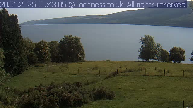 A grassy field with a wire fence overlooks a calm lake and distant mountains under a cloudy sky.