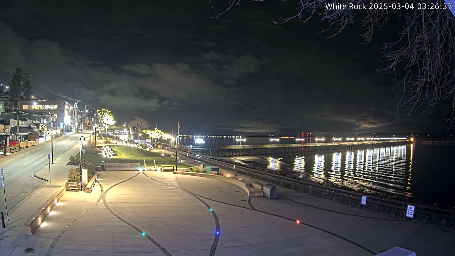 A nighttime view of a waterfront promenade with a long pier extending into calm water under a cloudy sky.