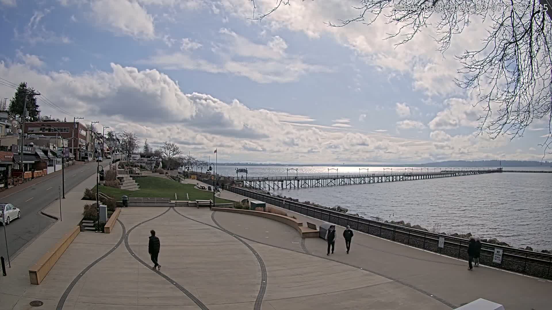 A long pier extends into a calm, partly cloudy body of water, with a paved waterfront area and a few pedestrians visible, along with buildings and a street in the background.