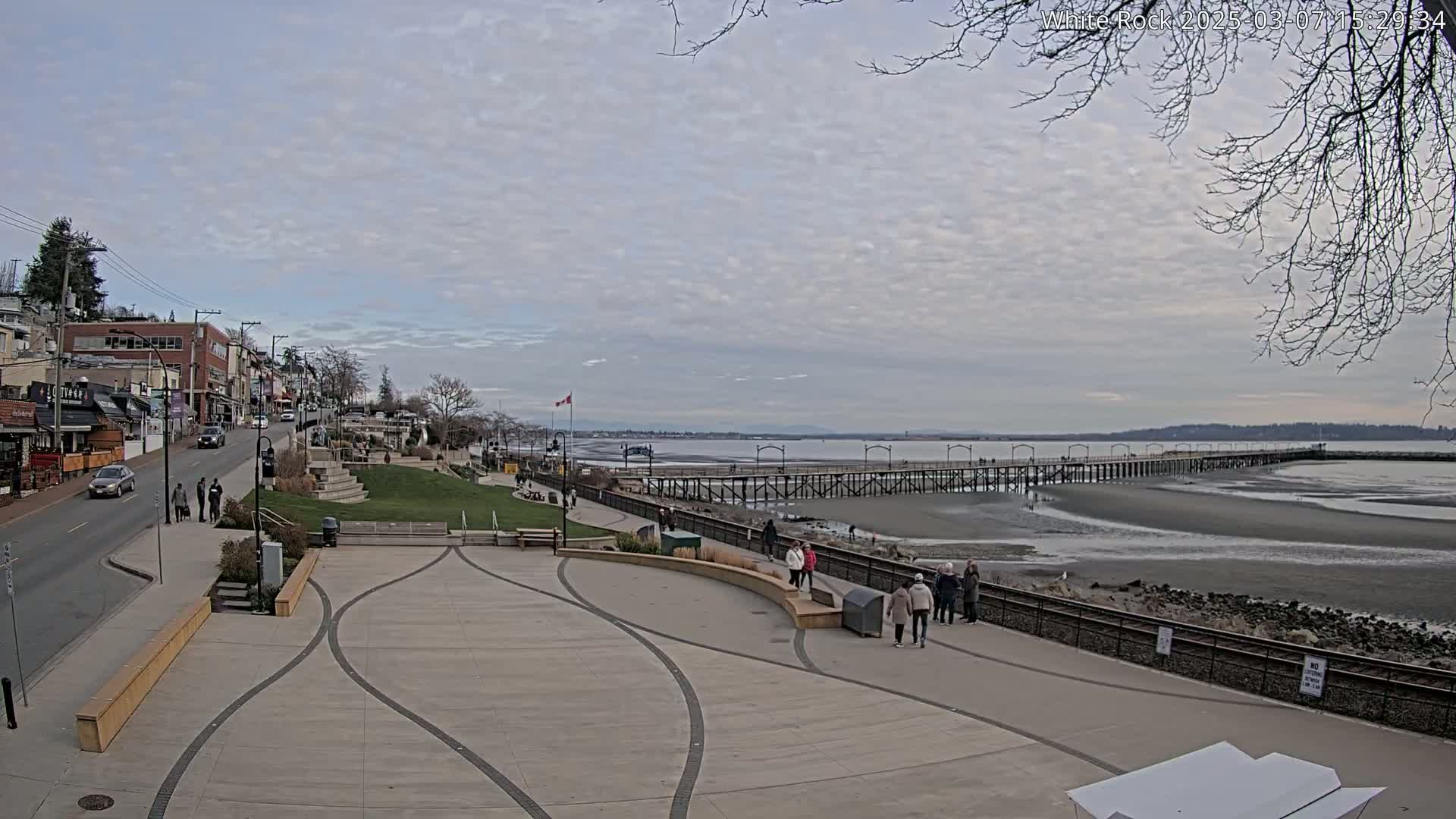 A paved plaza overlooks a long pier extending into a calm, overcast bay, with a street lined with buildings visible to the left.