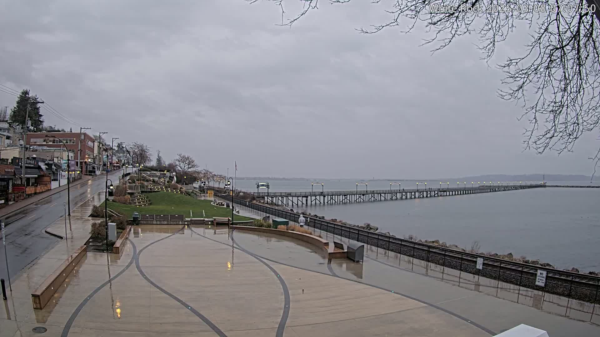 A wet, paved waterfront area with a long pier extending into a calm gray sea under an overcast sky.