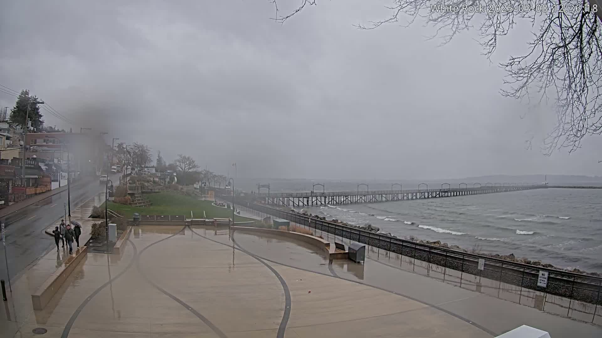 A long pier extends into a choppy gray sea under a cloudy, overcast sky, with a wet paved area and a few pedestrians in the foreground.