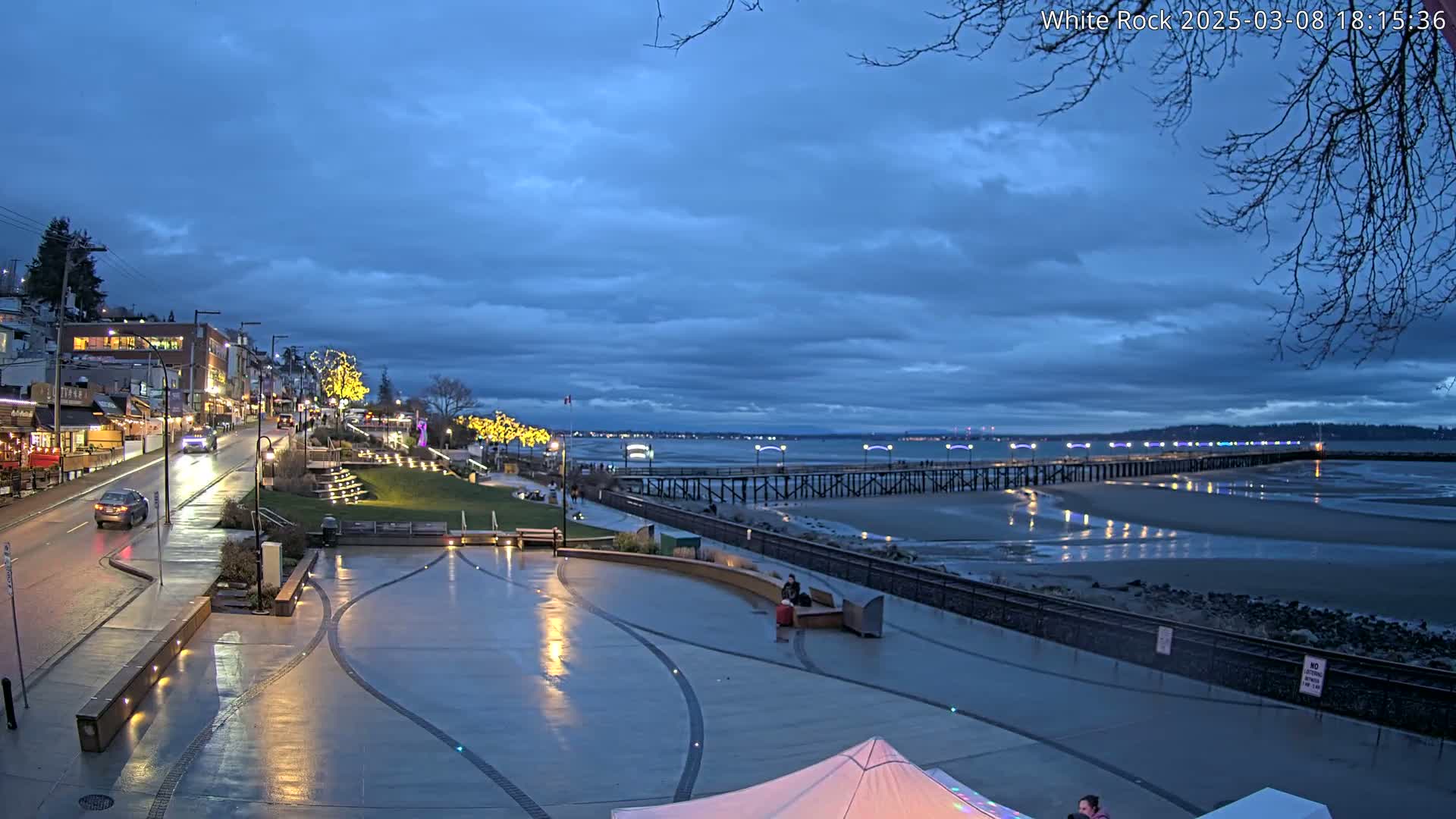 A wet, paved waterfront plaza at twilight, with buildings and a long pier extending into a calm, shallow bay under a partly cloudy sky.